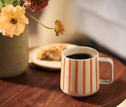 Eine weiße Tasse mit orangefarbenen Streifen, gefüllt mit Kaffee, steht auf einem Holztisch neben einer olivgrünen Vase mit pfirsichfarbenen und rötlichen Blumen. Ein Croissant ist im Hintergrund.