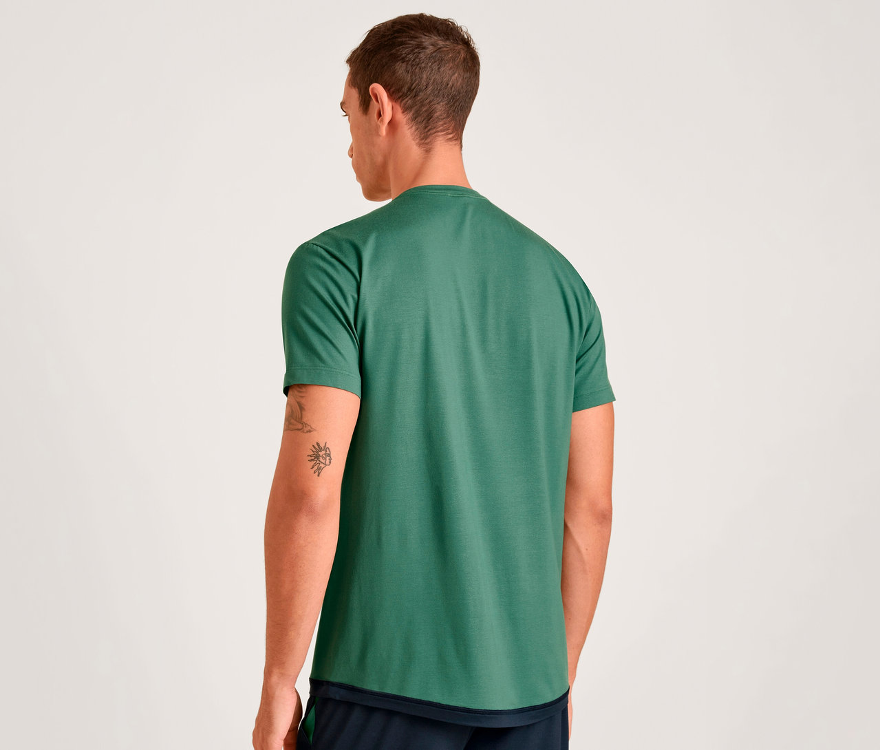 Mann von hinten gesehen, trägt ein grünes Kurzarm-T-Shirt und dunkle Shorts. Er hat Tätowierungen am rechten Arm.
