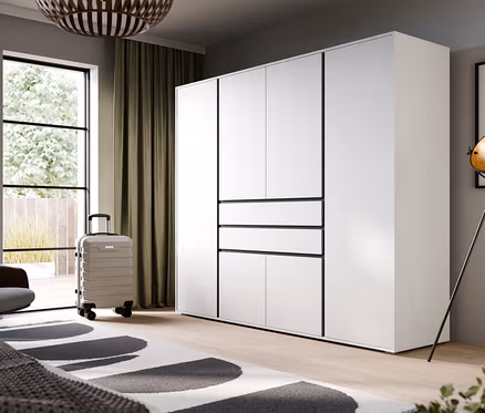 Modernes Schlafzimmer mit großem weißem Kleiderschrank mit schwarzen Akzenten, einem Fenster mit olivgrünen Vorhängen, einem grauen Sessel, einem hellgrauen Rollkoffer und einem weißen Teppich mit schwarzen Mustern.
