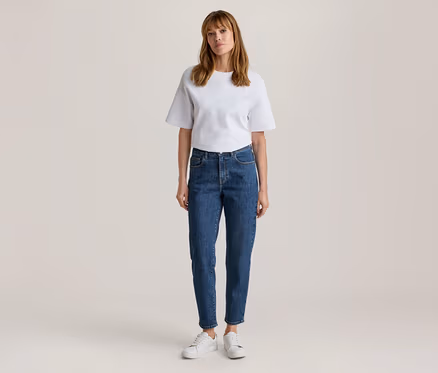 Frau steht, trägt ein weißes Kurzarm-T-Shirt, blaue Jeans und weiße Turnschuhe.