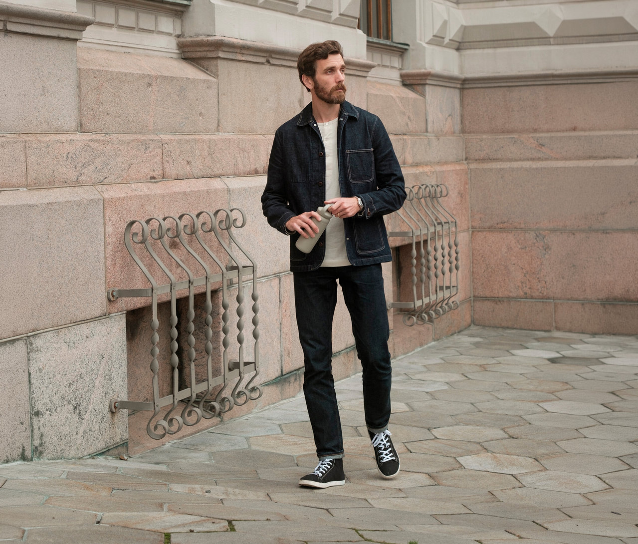 Mann mit Bart in Jeansjacke und Jeans, der einen beigen KINTO Tumbler »Day Off« hält und nach rechts blickt.