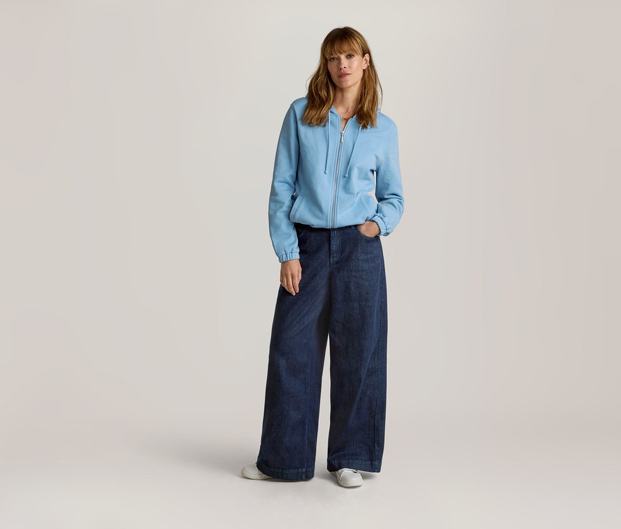 Frau mit langen, hellbraunen Haaren trägt eine hellblaue Sweatjacke mit Reißverschluss und weite, dunkelblaue Jeans, eine Hand in der Tasche.