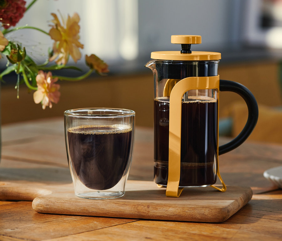 Ein doppelwandiges Glas mit schwarzem Kaffee und eine gelbe French Press, gefüllt mit Kaffee, stehen auf einem Holzbrett. Im Hintergrund sind eine Blumenvase und ein Teller mit einem Keks zu sehen.