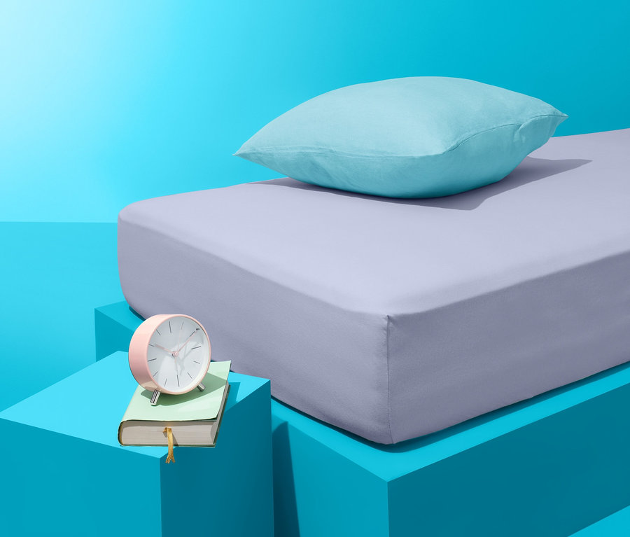 Auf blauem Hintergrund befinden sich ein Bett mit lila Bettlaken und ein blaues Kissen. Neben dem Bett steht ein blauer Tisch mit einem Buch und einem rosa Wecker.