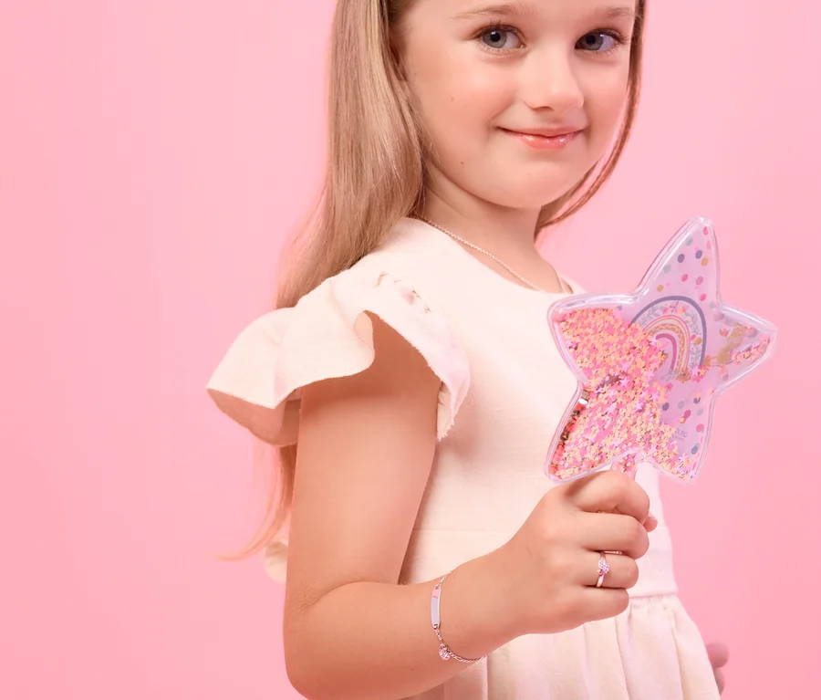 Ein lächelndes Mädchen mit blonden Haaren hält einen sternförmigen Zauberstab. Sie trägt ein Amor 925 Silber Kinder-Armband mit Herz-Anhänger und Plättchen am linken Handgelenk und einen pinken Ring am linken Ringfinger.