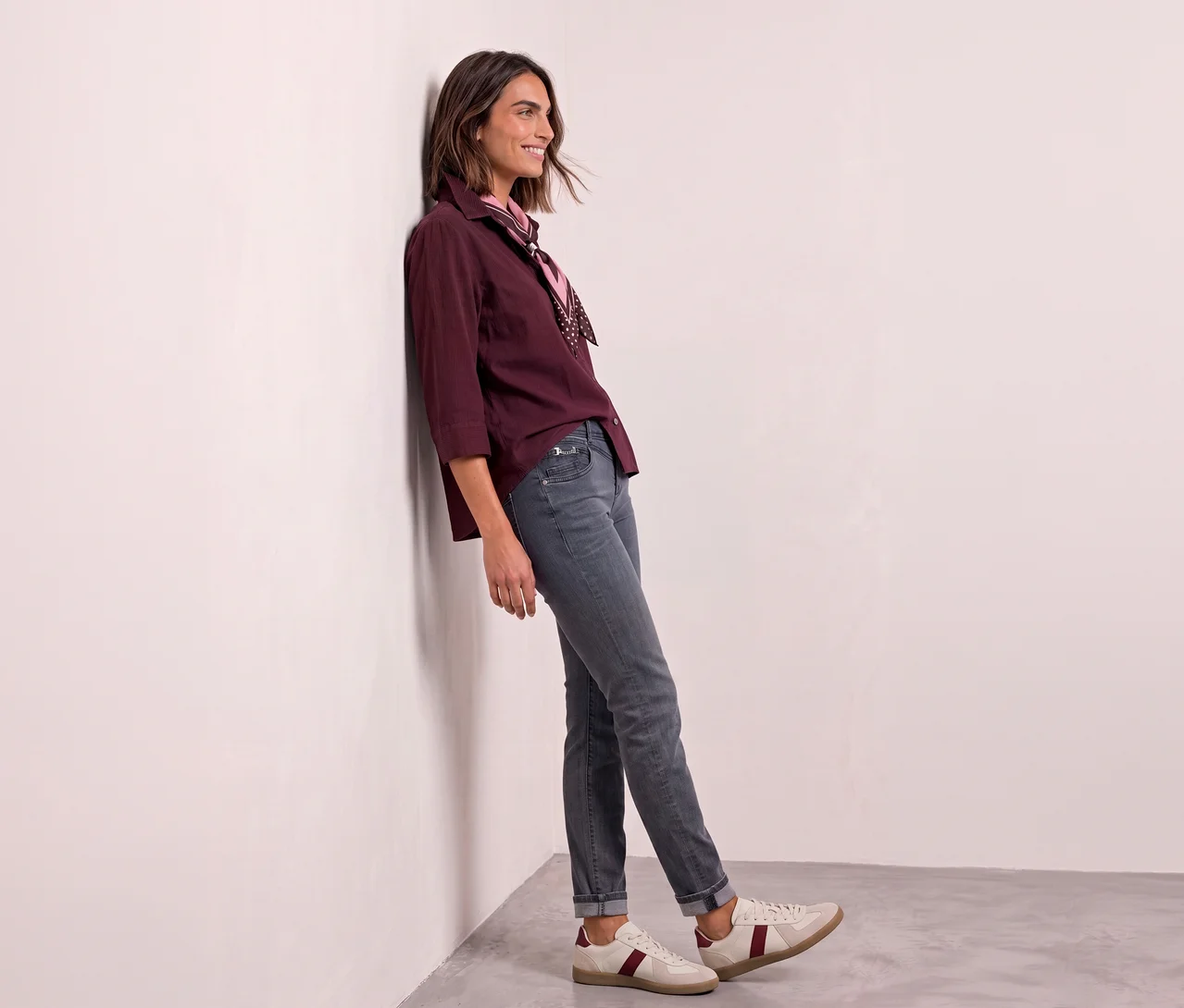 Eine lächelnde Frau lehnt an einer weißen Wand und trägt eine bordeauxrote Bluse, ein rosa Halstuch und eine graue Street One Jeans.