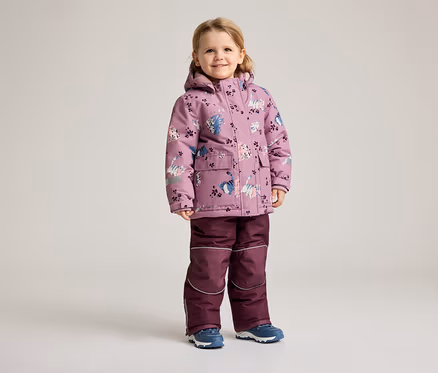 Ein kleines Mädchen steht in einem lila Wintermantel mit Katzenmuster und einer passenden Hose vor einem hellen Hintergrund.