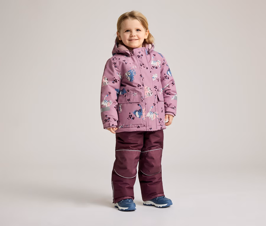 Ein kleines Mädchen steht in einem lila Wintermantel mit Katzenmuster und einer passenden Hose vor einem hellen Hintergrund.