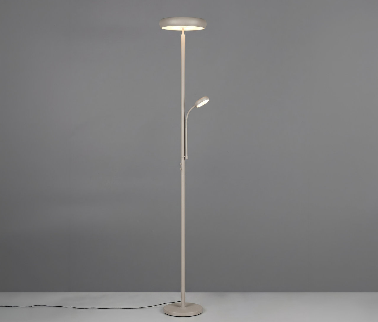 Eine hellgraue Stehleuchte mit zwei Lichtern. Das Hauptlicht ist ein Deckenfluter oben, und eine kleinere, verstellbare Leselampe ragt aus der Mitte des Ständers. Die Lampe steht auf einem hellen Boden vor einer grauen Wand, ein schwarzes Stromkabel ist auf dem Boden sichtbar.