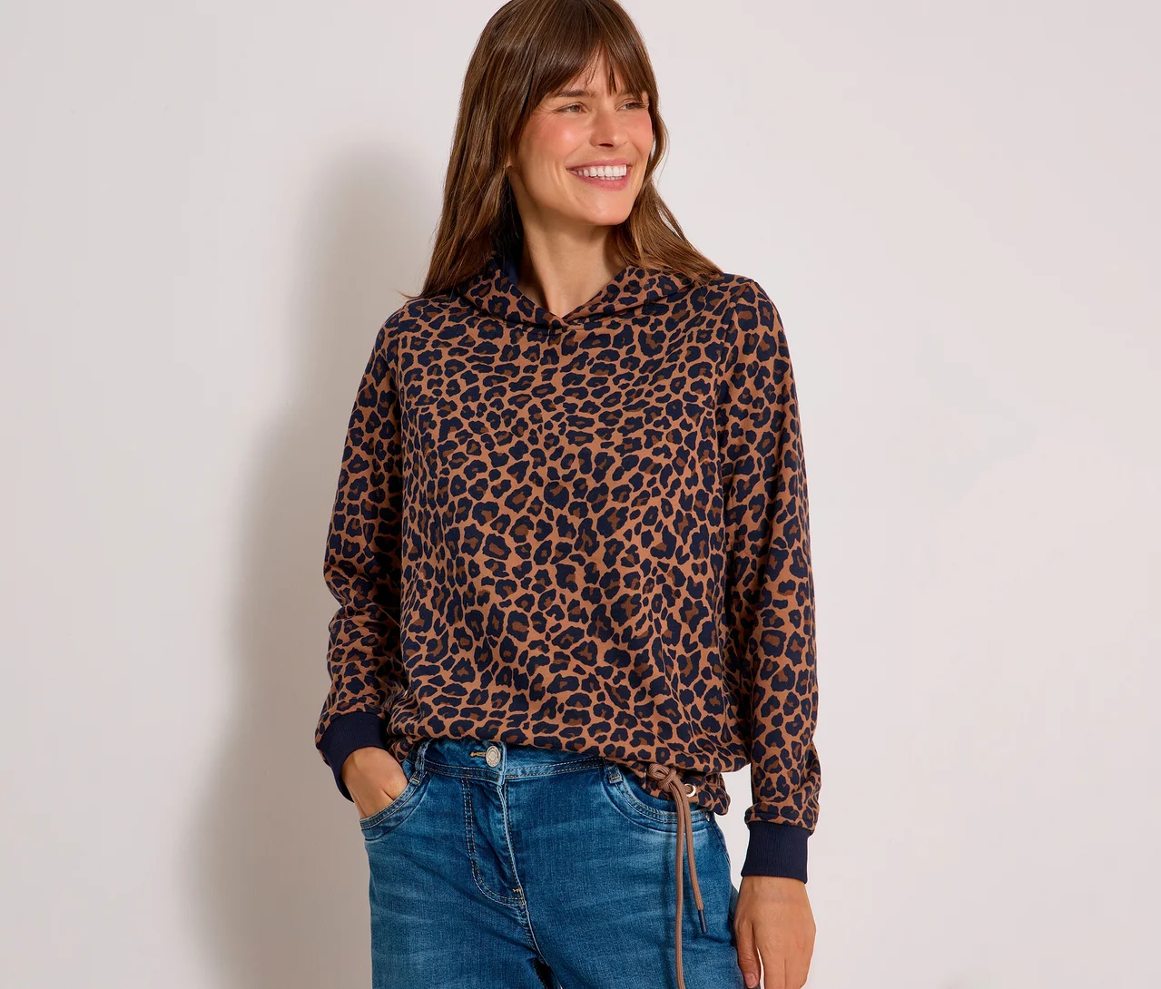 Lachende Frau trägt ein braun-dunkelblaues CECIL Sweatshirt mit Leopardenmuster und Kapuze sowie blaue Jeans, Hände in den Taschen.