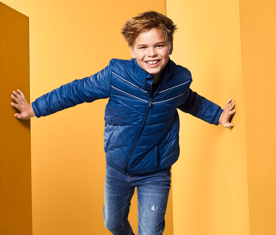 Ein Junge in einer blauen Steppjacke steht in der Ecke eines gelben Raumes.
