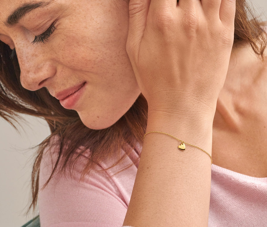 Nahaufnahme einer Frau mit braunen Haaren, die ein Gold-Armband mit Herzanhänger und einen Gold Ring V Zirkonia trägt.