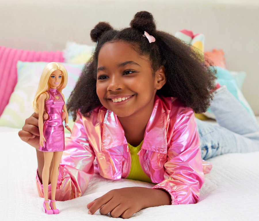 Lächelndes Mädchen liegt mit pinkfarbener Jacke auf dem Bett und hält eine Barbie-Puppe mit pinkfarbenem Kleid.