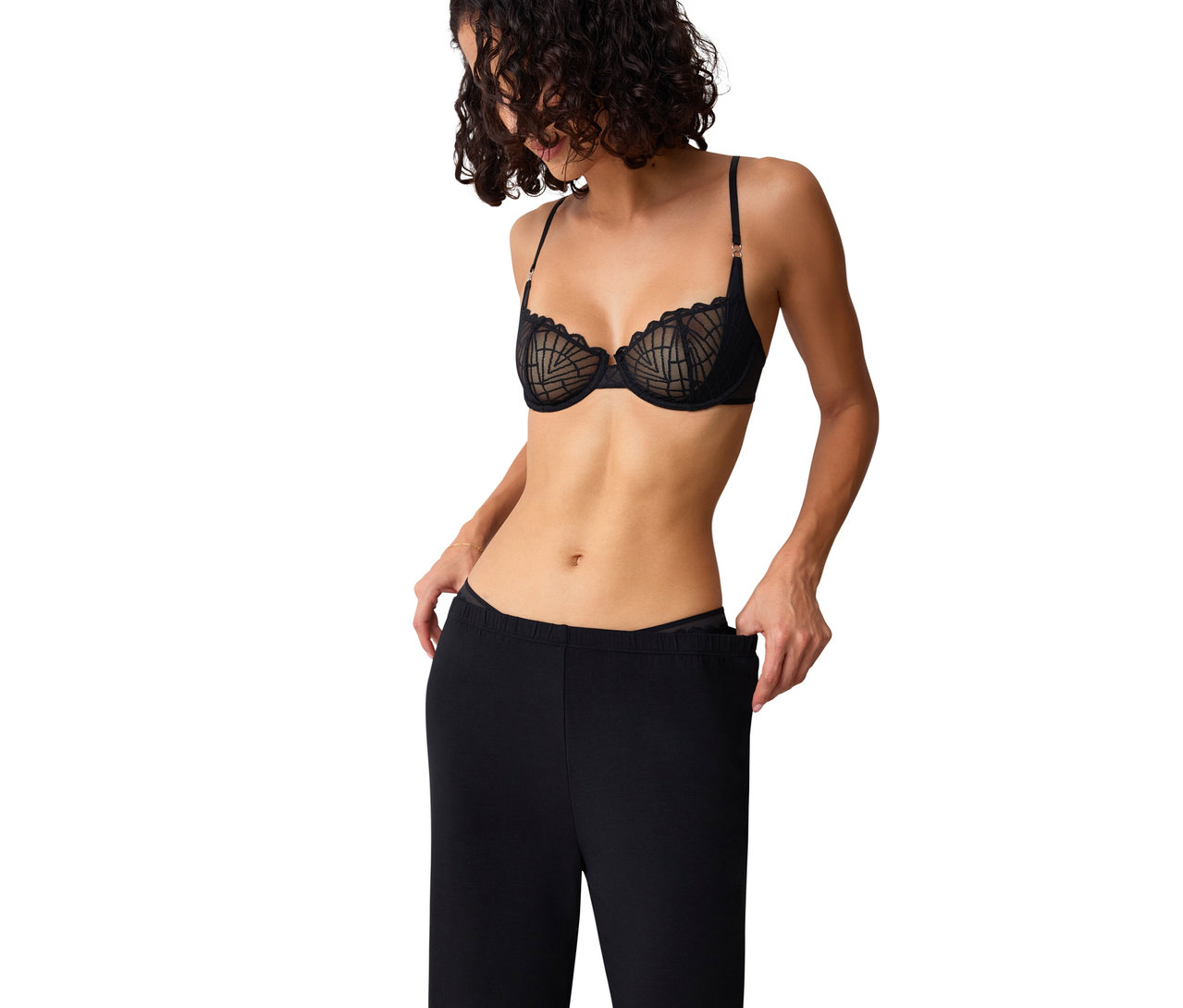 Frau mit lockigem Haar trägt einen schwarzen Triumph That Night in Vegas WH Bügel-BH mit geometrischem Muster und zieht den Bund ihrer schwarzen Hose herunter.
