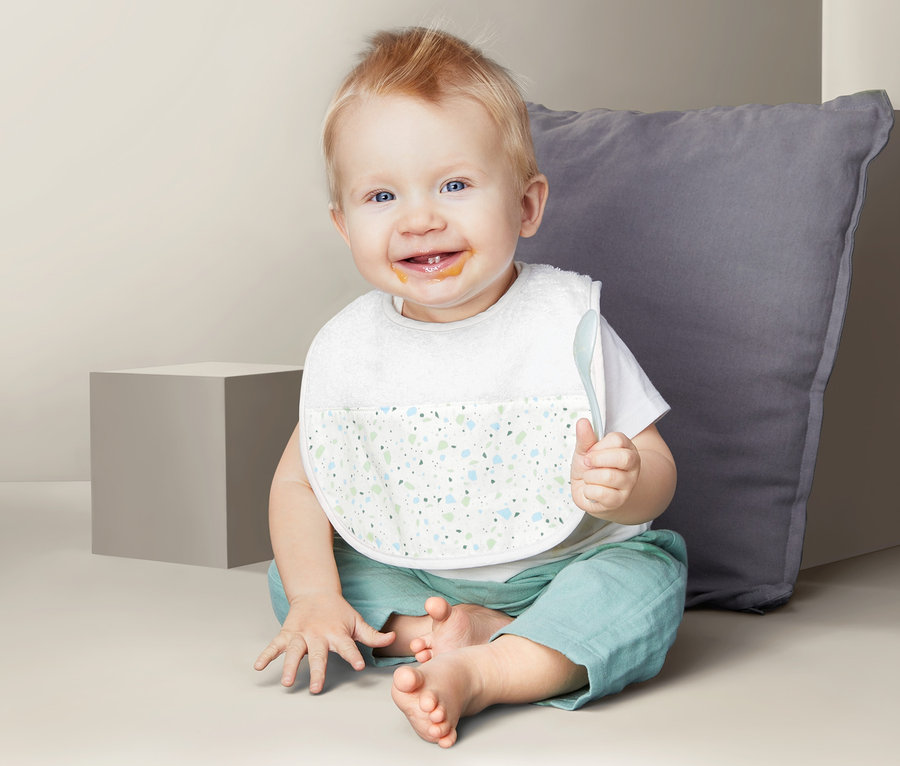 Lächelnder Säugling sitzt mit 2 Baby-Lätzchen und einem Löffel in der Hand auf dem Boden.