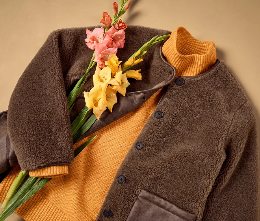 Brauner flauschiger Merino-Cardigan liegt offen über einem orangefarbenen Rippstrickpullover, darauf rosa und gelbe Gladiolen.