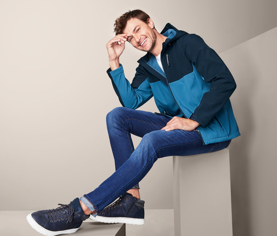 Mann sitzt und lächelt, trägt eine blaue Softshelljacke.