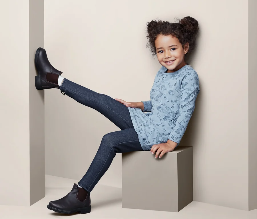 Lächelndes kleines Mädchen sitzt auf einem Würfel und trägt Thermotreggings und Stiefeletten.