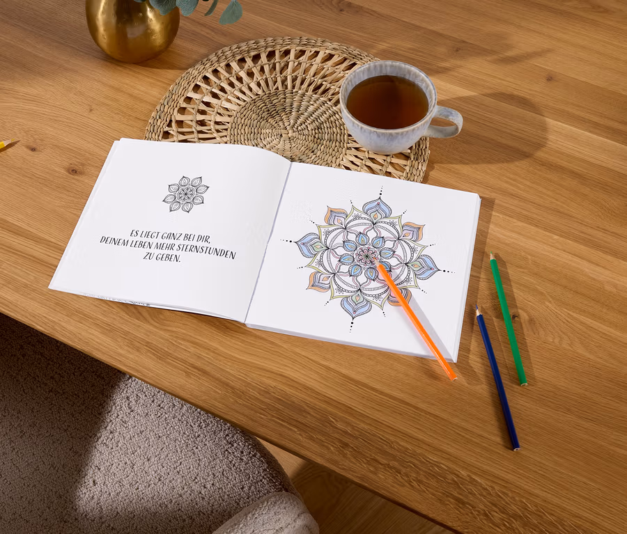 Ein geöffnetes Malbuch mit einem Mandala liegt auf einem Holztisch neben einer Tasse Tee und bunten Stiften.