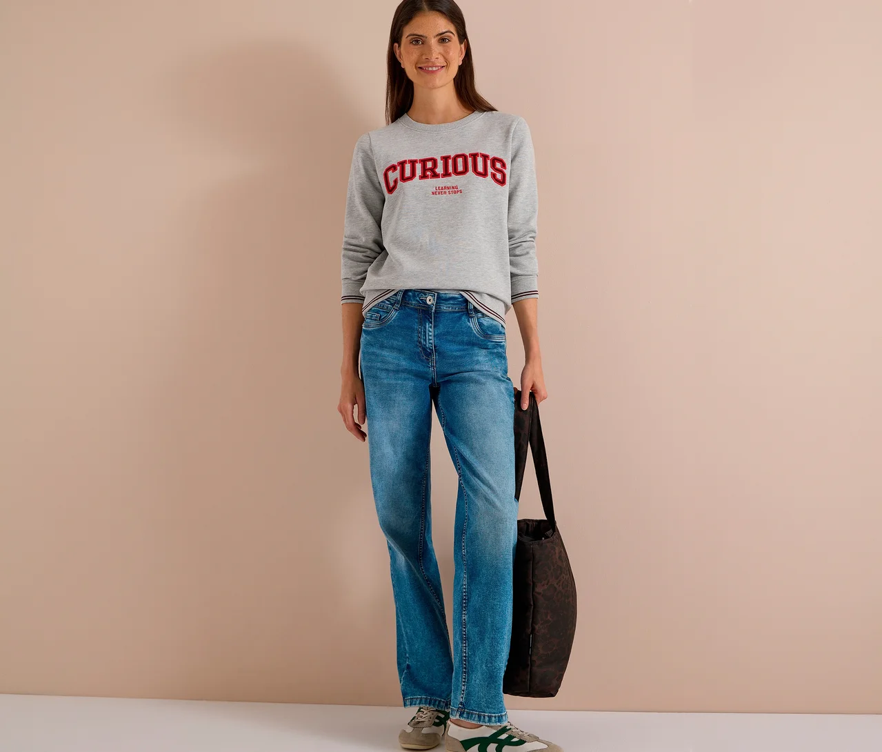 Eine lächelnde Frau steht in einem grauen CECIL Sweatshirt mit rotem "CURIOUS" Aufdruck, blauen Jeans und beigen Sneakern, eine dunkle Tasche haltend.