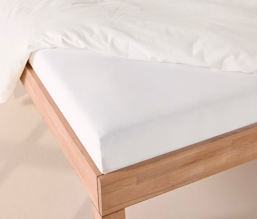 Weißes Jersey-Spannbettlaken auf einem Holzbett, mit einer weißen Bettdecke darüber.
