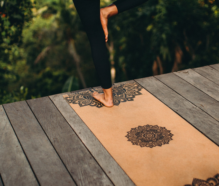 Frau steht auf einem Bein auf einer Yogamatte mit Mandala-Muster auf einem Holzdeck.