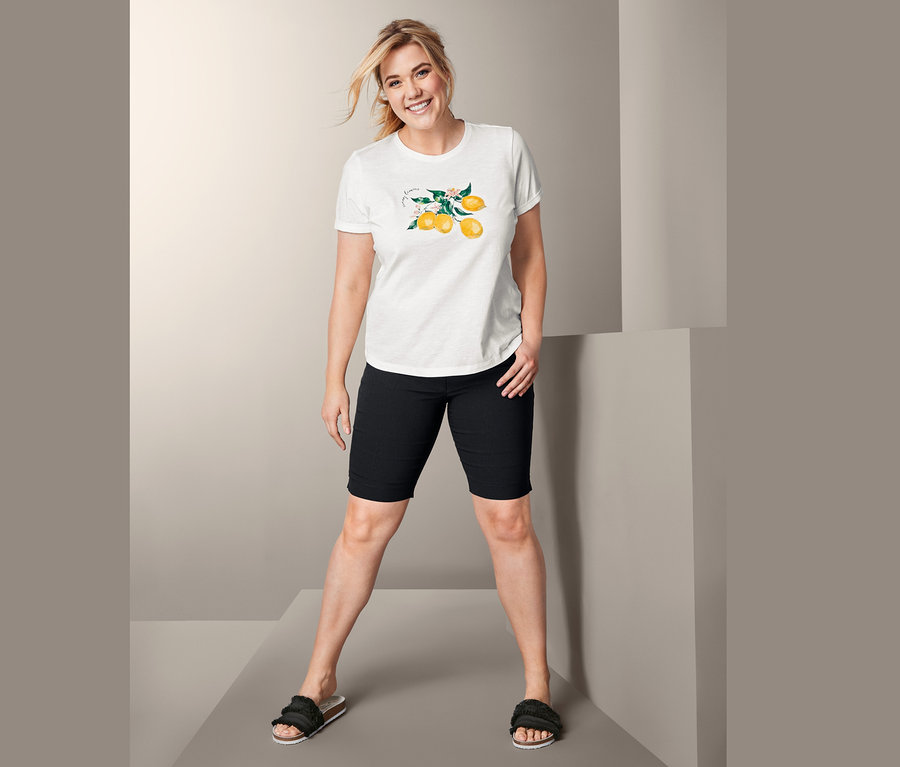 Frau posiert in bedrucktem Kurzarmshirt, schwarzen Shorts und Pantoletten.