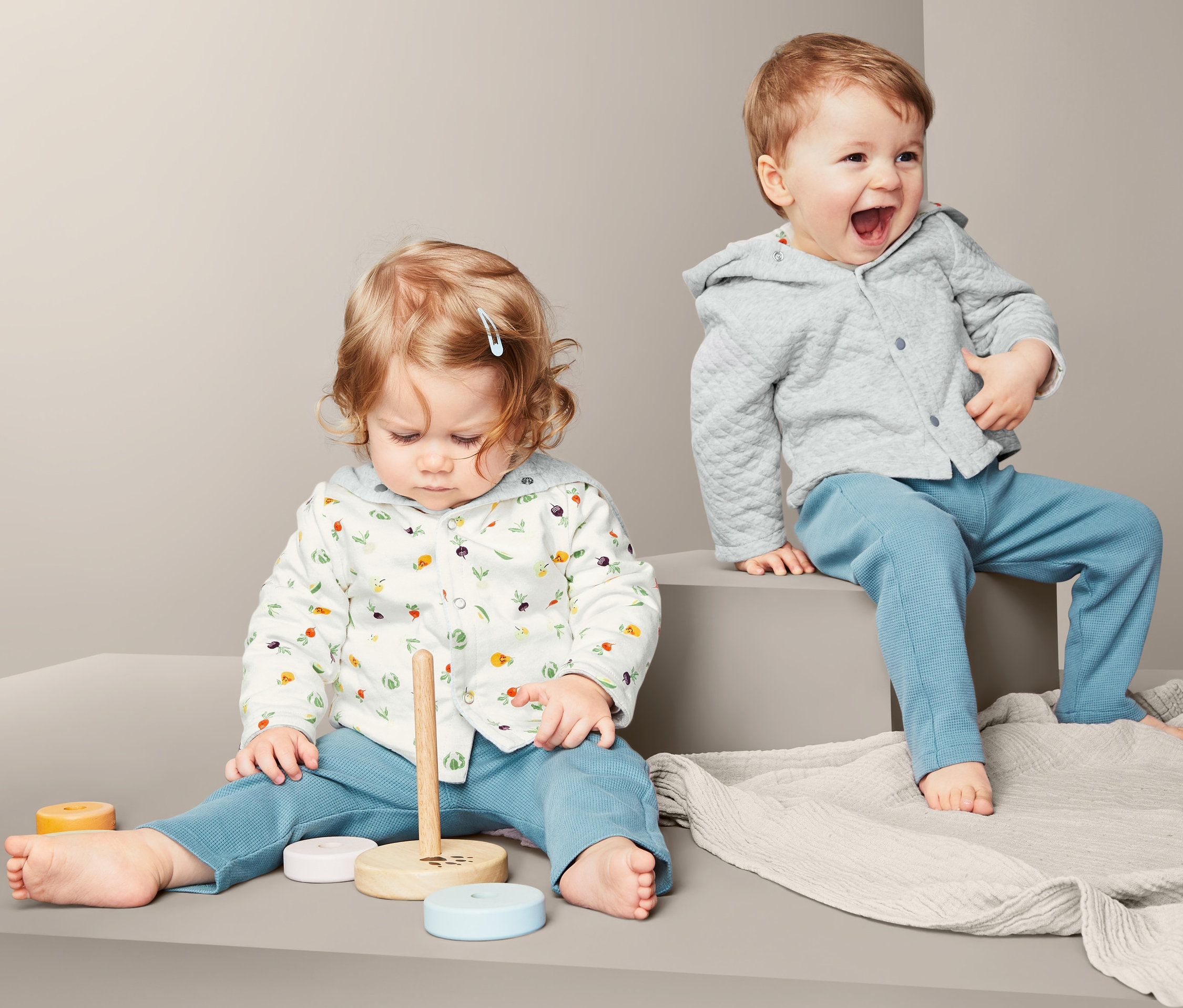 Zwei Kleinkinder sitzen und spielen. Eines trägt eine gesteppte Jerseyjacke und lacht.