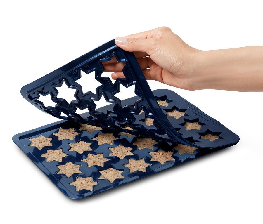 Eine Hand entfernt eine Zimtstern-Backmatte aus Silikon von einem Backblech mit sternförmigem Teig.