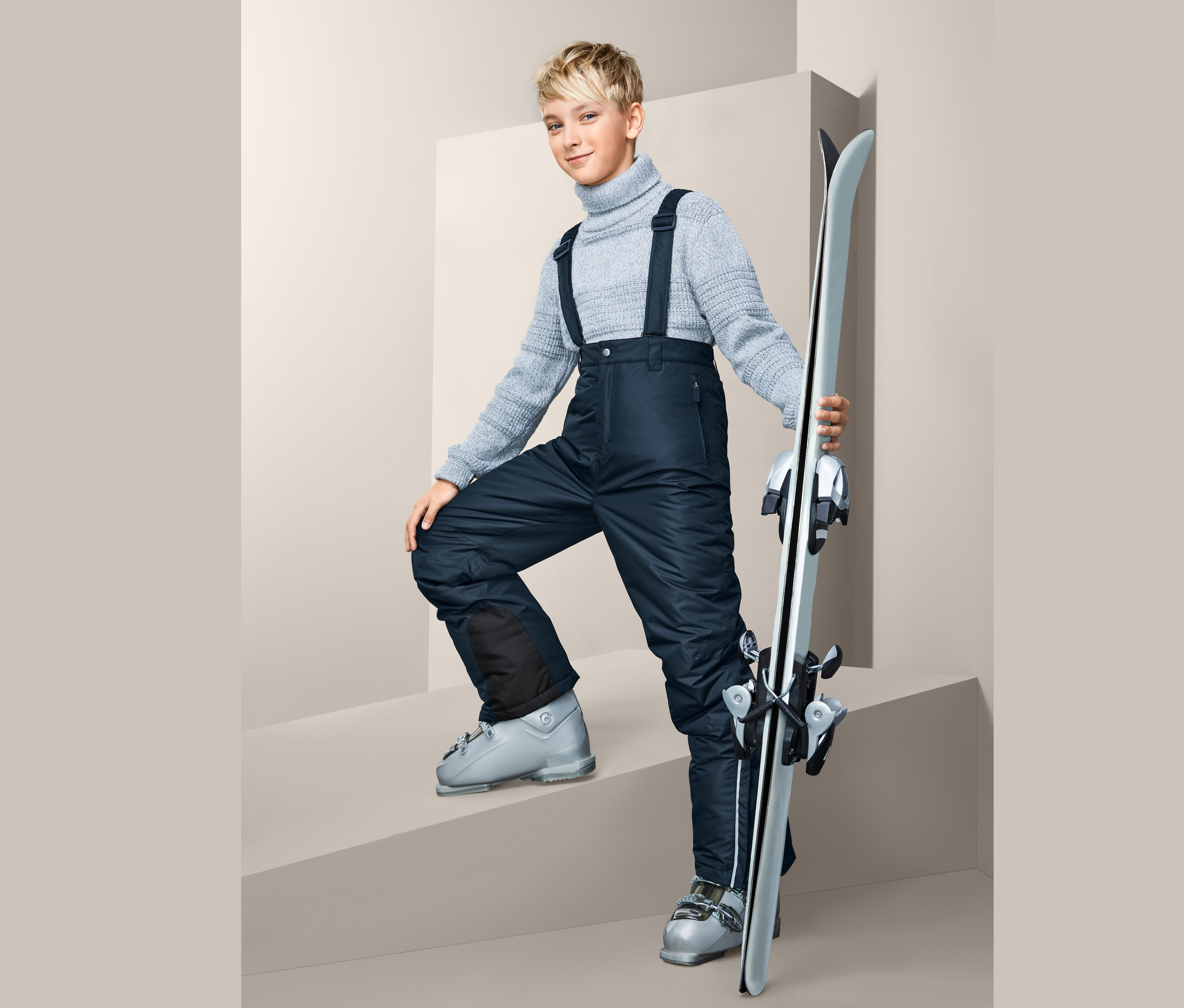 Junge mit grauem Pullover und Skihose steht auf einer Treppe und hält Ski.