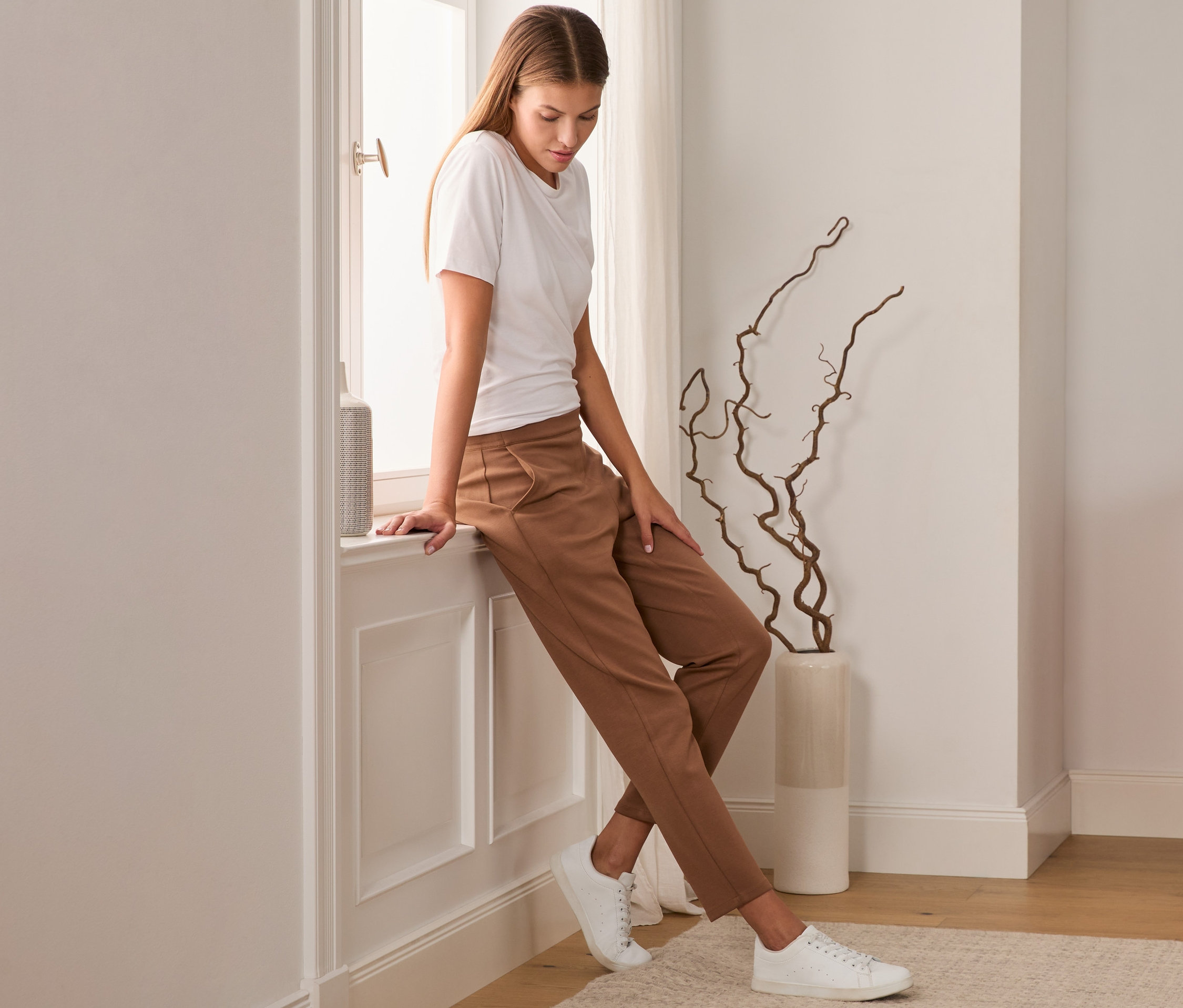 Eine Frau sitzt mit braunen Joggpants auf einem Fensterbrett.