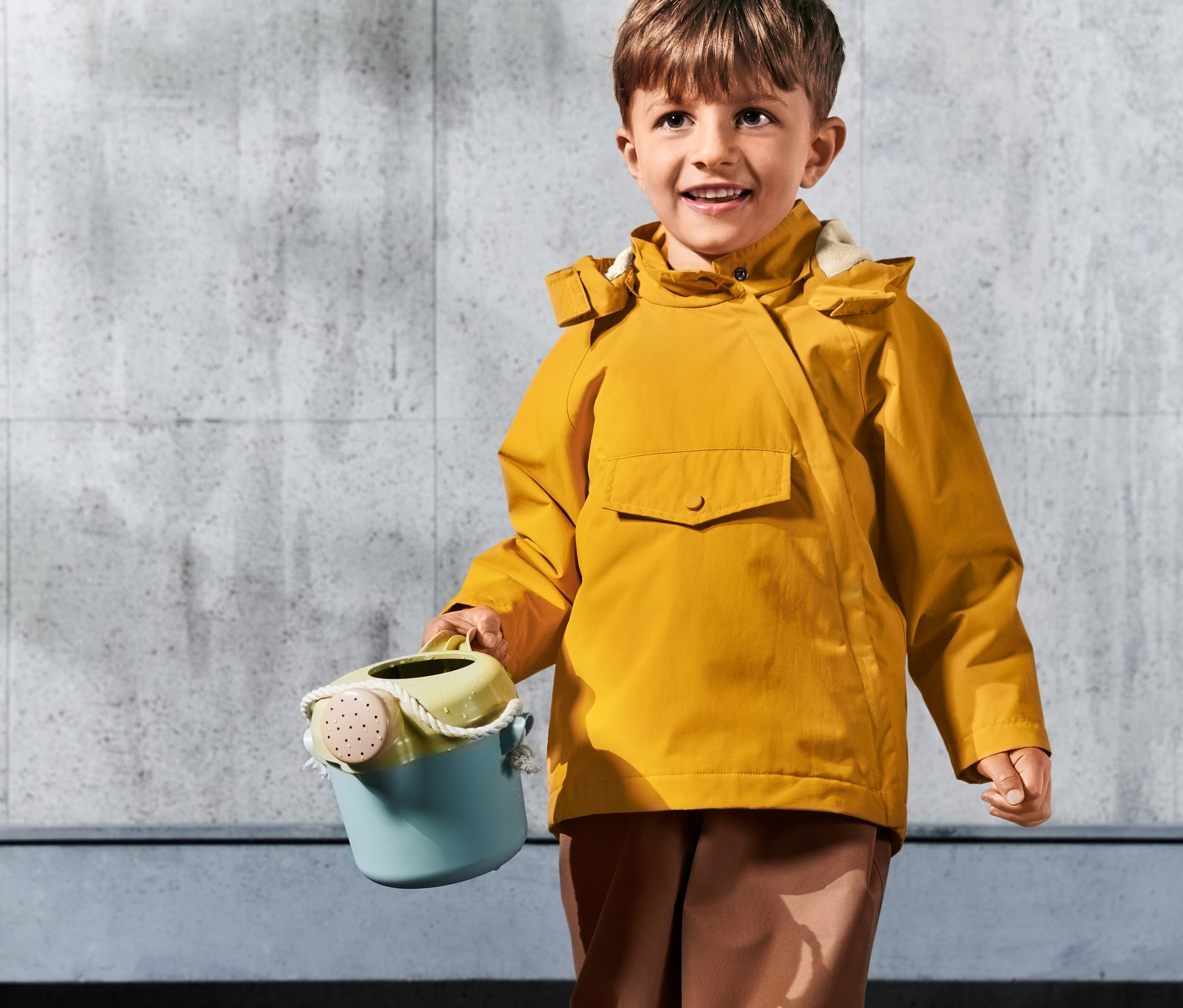 Junge hält eine blaugrüne Gießkanne und trägt eine gelbe Thermo-Regenjacke.