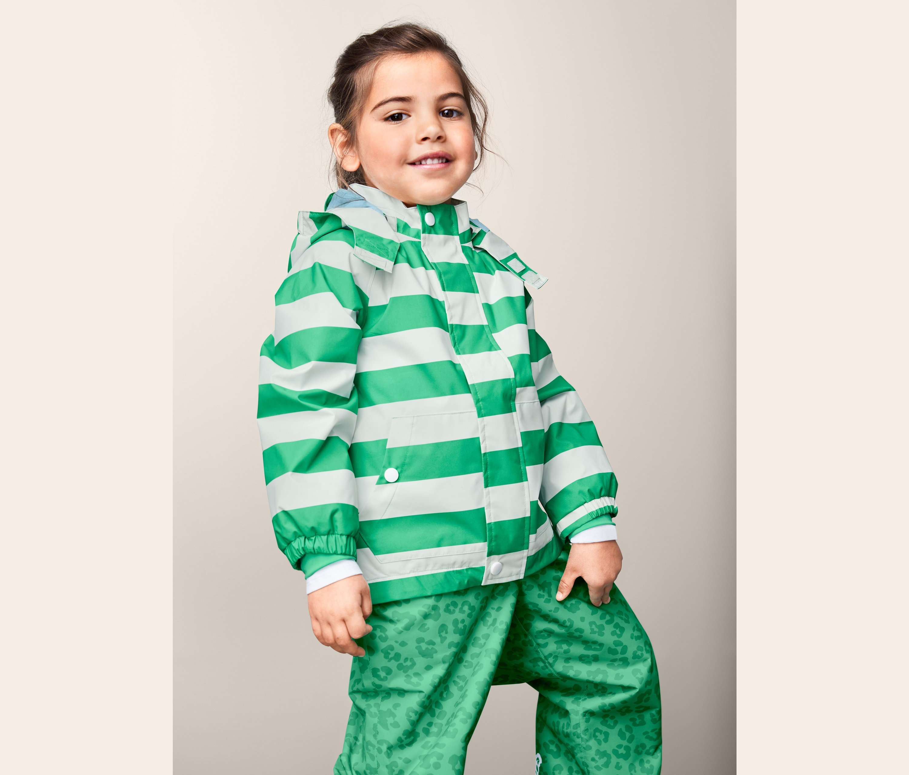 Mädchen posiert in einer Regenjacke aus recyceltem Material mit grün-weißen Streifen und einer grünen Hose mit Print.