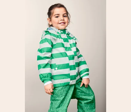 Mädchen posiert in einer Regenjacke aus recyceltem Material mit grün-weißen Streifen und einer grünen Hose mit Print.