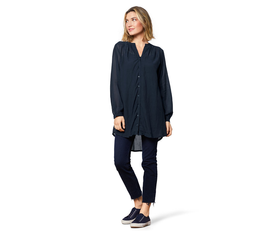 Frau stehend, trägt eine Tunika, Jeans und Turnschuhe.