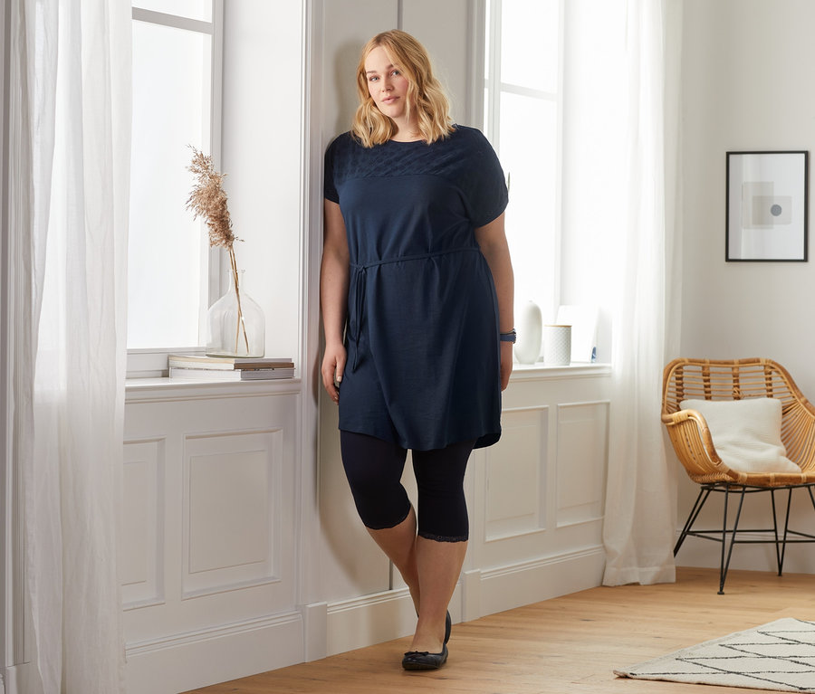 Blonde Frau lehnt am Fenster und trägt ein dunkelblaues Jerseykleid mit Lochstickerei.