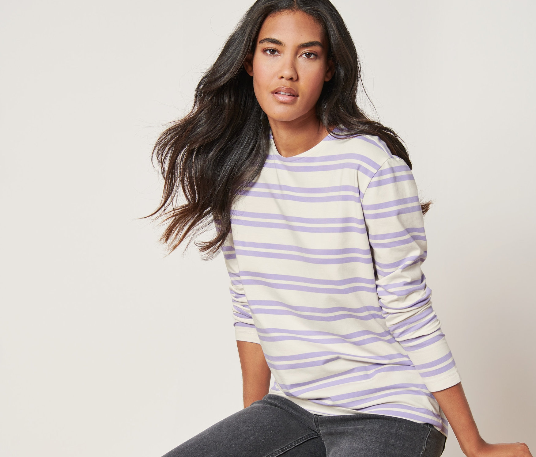 Frau sitzt auf einem Stuhl und trägt ein lila-cremefarben gestreiftes Langarmshirt.