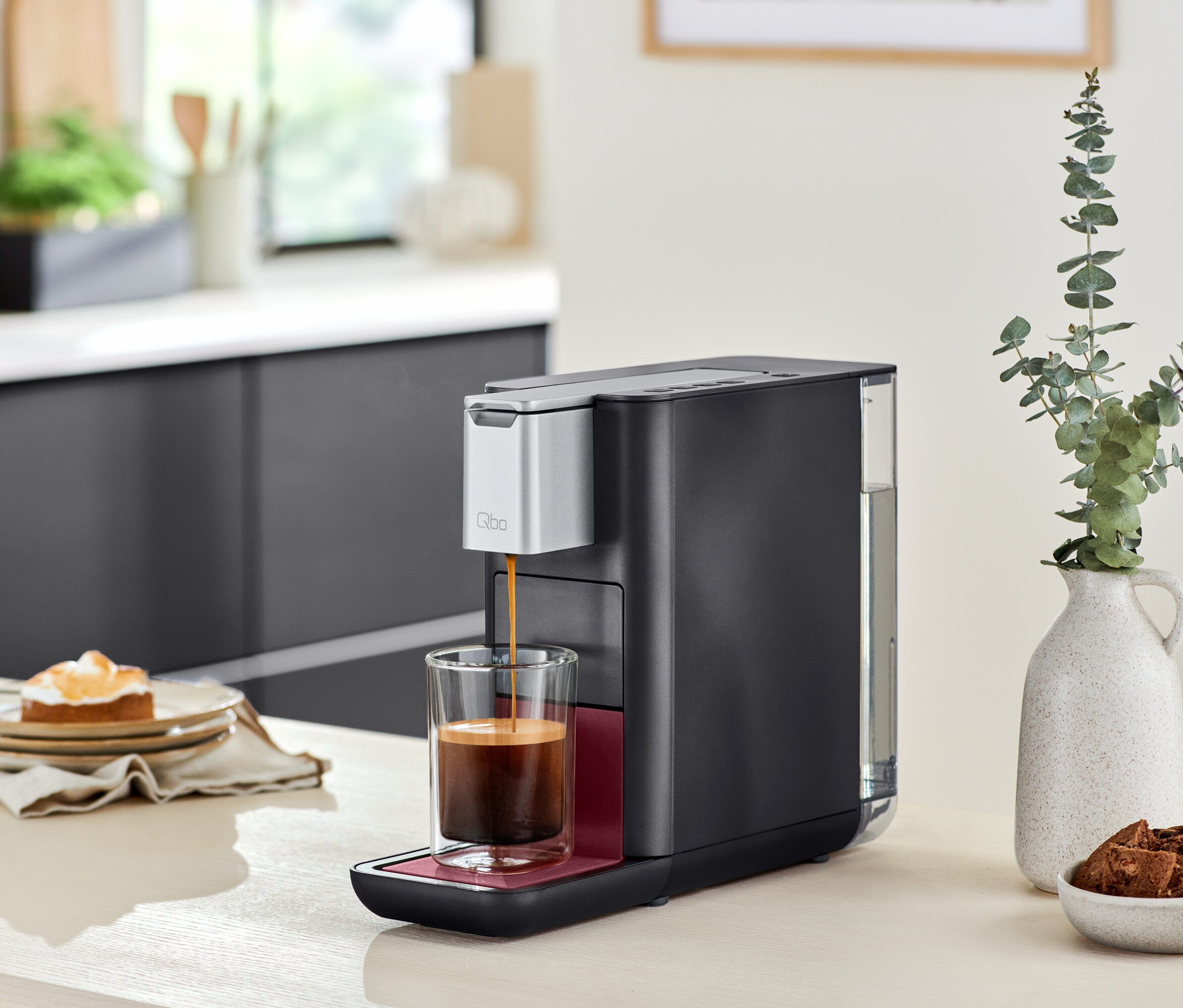 Qbo ESSENTIAL Dark Berry Kaffeemaschine gießt Kaffee in ein Glas, neben einem Teller mit Kuchen und einer Vase mit Blumen.