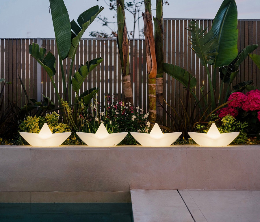 Vier Goodnight Light LED-Leuchten »Boot« stehen auf einer Mauer am Pool, im Hintergrund Pflanzen und ein Holzzaun.
