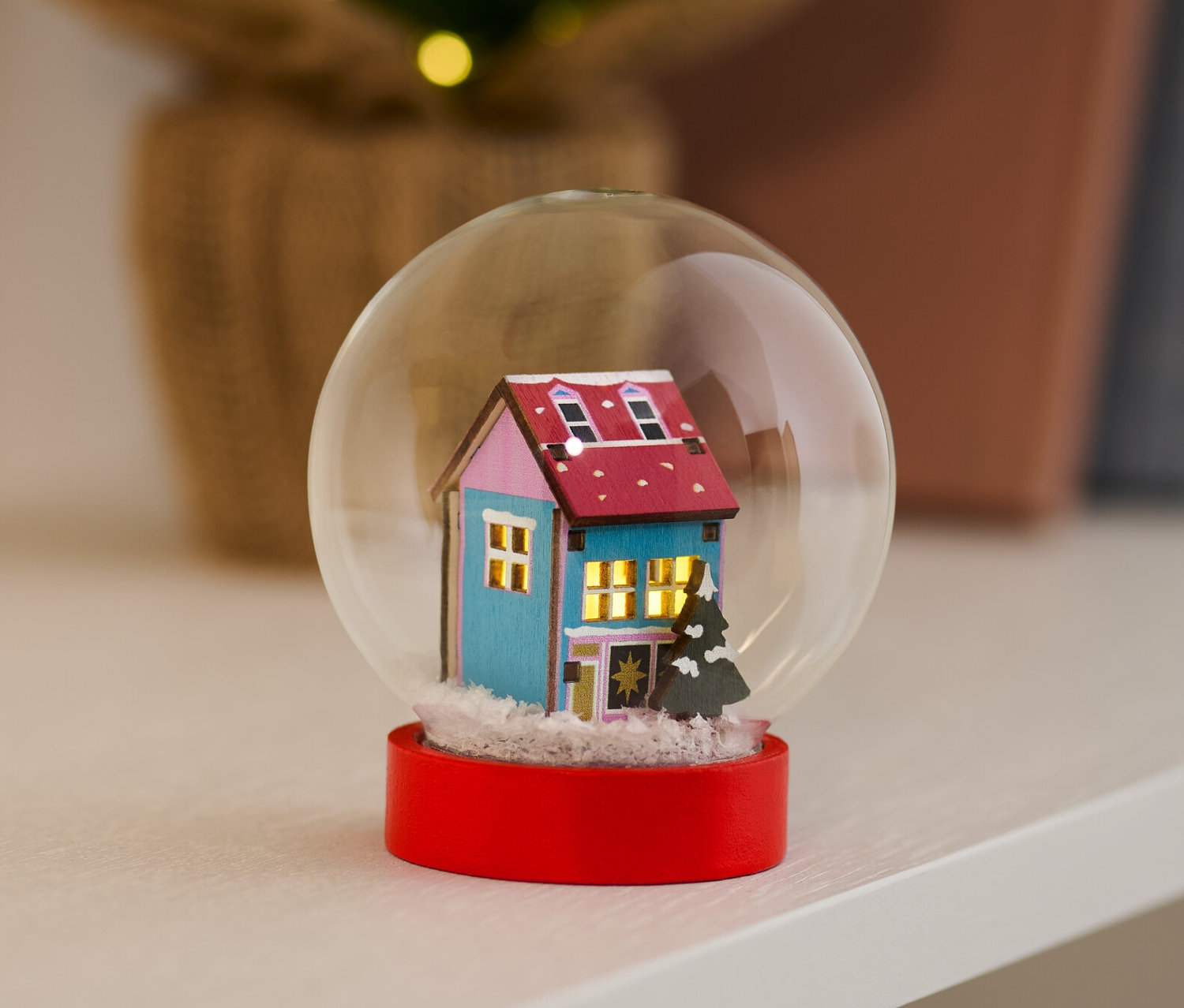 LED-Schneekugel mit blauem Haus und rotem Sockel steht auf einem Regal.