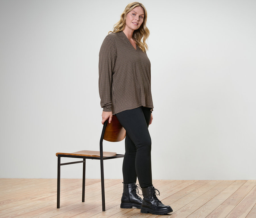 Frau posiert in bedrucktem Blusenshirt mit V-Ausschnitt und Heavy-Leggings neben einem Stuhl.