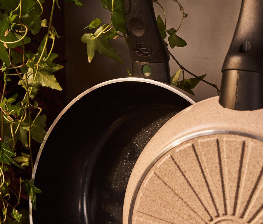 Zwei Pfannen hängen an Haken neben einer Pflanze. Eine davon ist eine sandfarbene BALLARINI Aluminium-Bratpfanne, 28 cm.