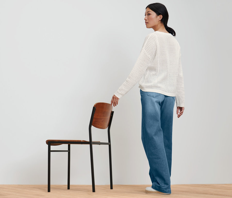 Frau posiert in Pointellestrickpullover und weiter Jeans neben einem Stuhl.