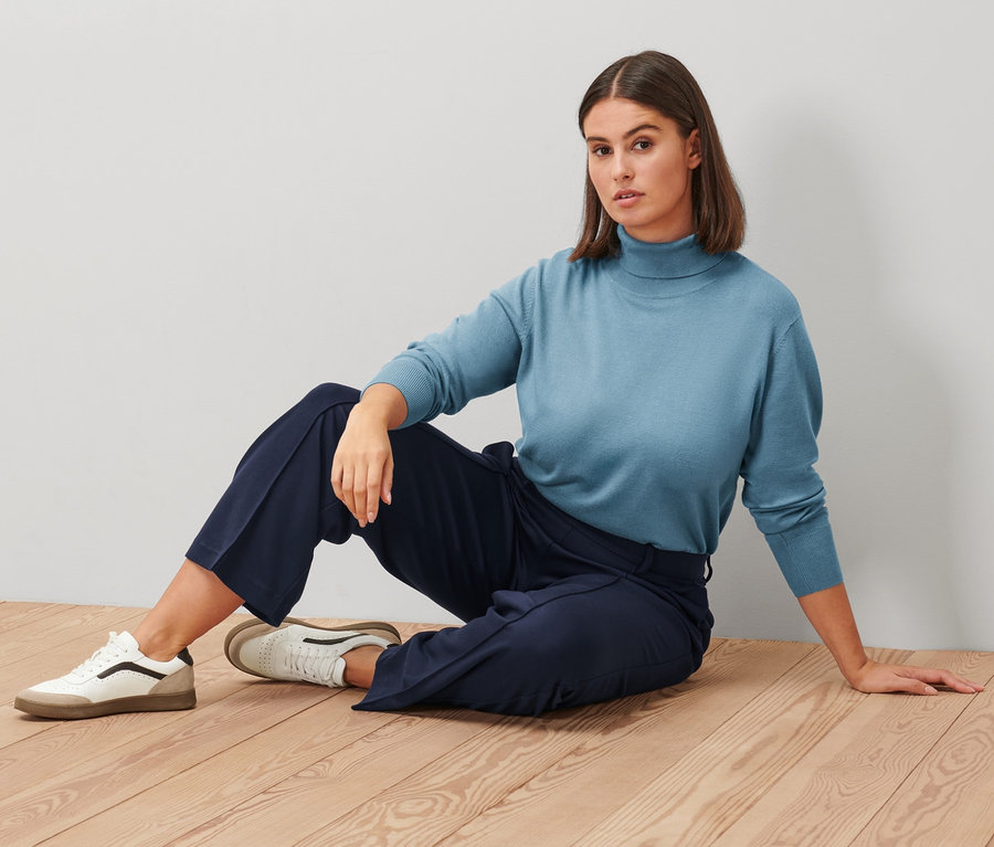 Frau sitzt auf einem Holzboden und trägt einen rauchblauen Feinstrick-Rollkragenpullover, eine dunkelblaue Jerseyhose mit Biese und Leder-Sneaker.