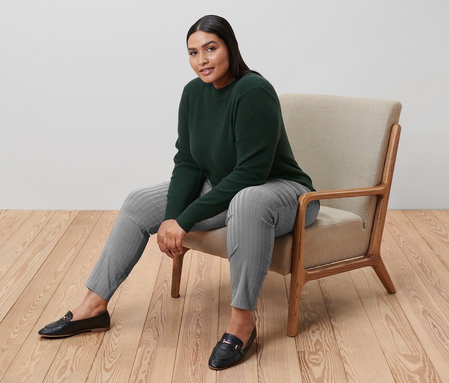 Frau sitzt im Sessel und trägt einen grünen Feinstrickpullover und eine graue bedruckte Stretchhose.