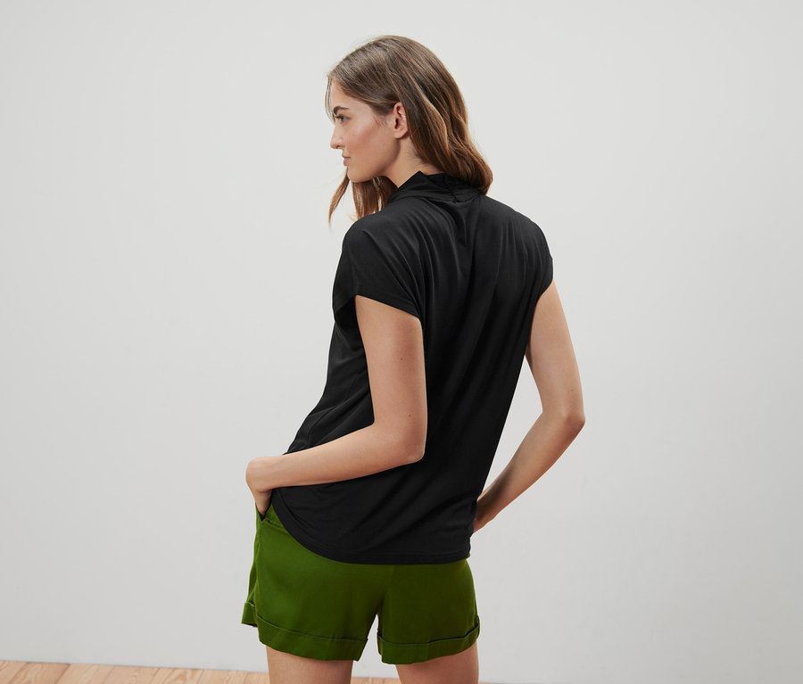 Frau posiert mit dem Rücken zur Kamera und trägt ein schwarzes Blusenshirt mit V-Ausschnitt und grüne Shorts.