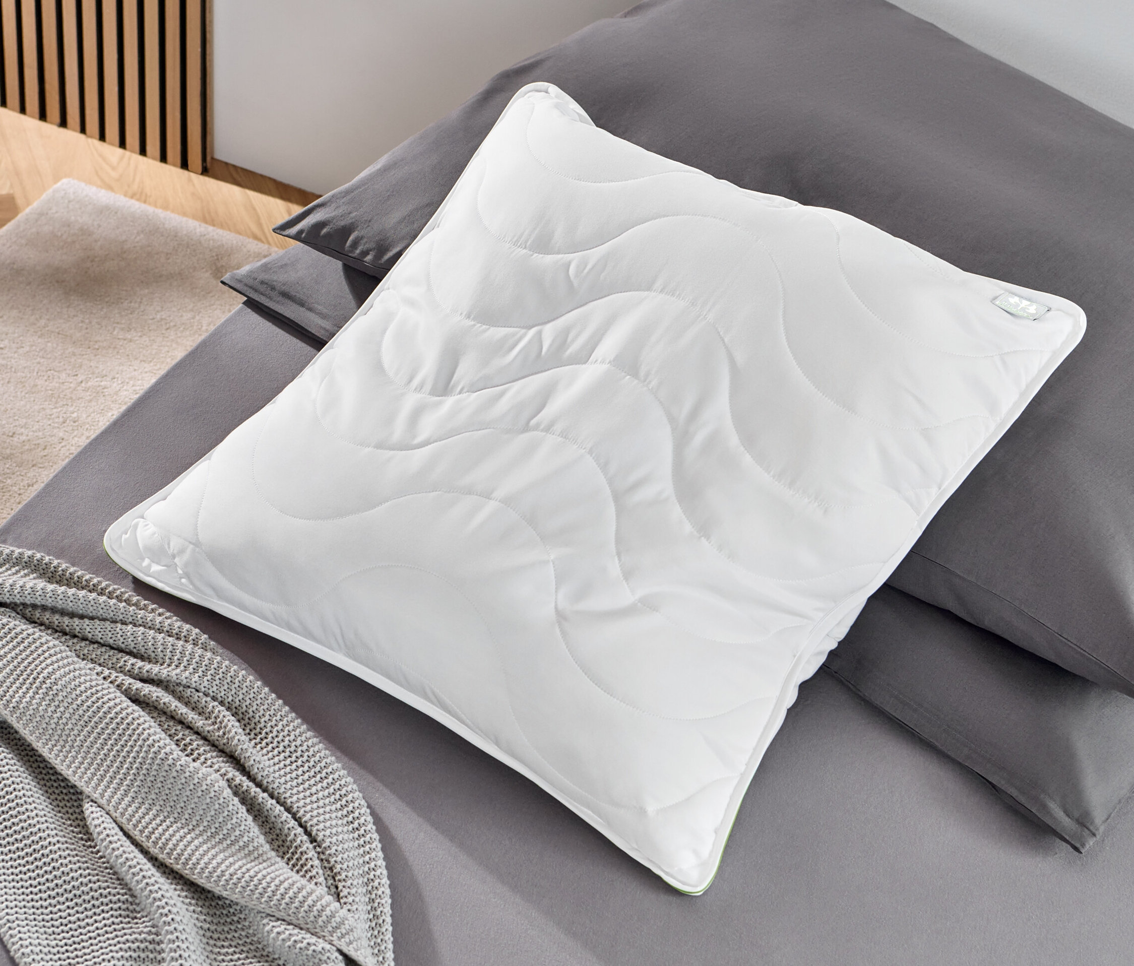 Ein weißes irisette® greenline Kopfkissen, ca. 80 x 80 cm, liegt auf grauer Bettwäsche neben einer grauen Decke.