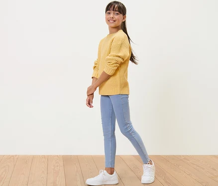 Mädchen posiert in Kinder-Chenille-Pullover, rapsgelb, hellblauen Jeans und weißen Turnschuhen.