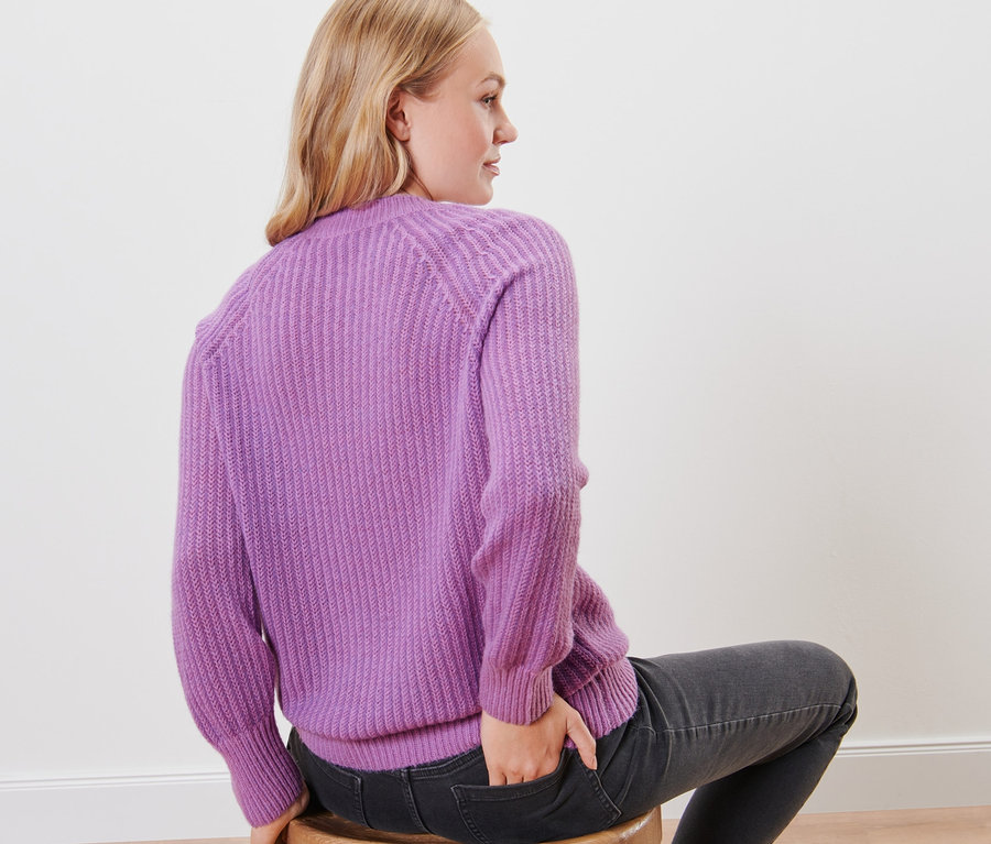 Frau sitzt auf einem Hocker mit dem Rücken zur Kamera und trägt einen fliederfarbenen Strickpullover.