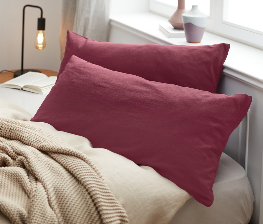 Zwei bordeauxrote Jersey-Kopfkissenbezüge liegen auf einem Bett mit einer beige Decke.
